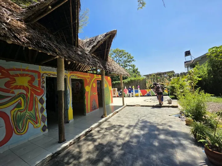 Thatched makuti roofs over a mural-painted walkway beside a sunny courtyard garden