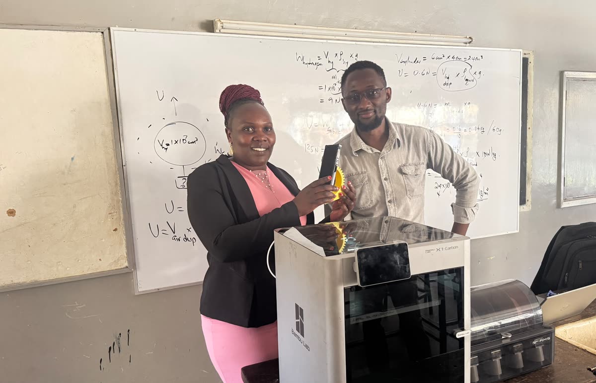 Two adults stand at a whiteboard covered in handwritten equations; one holds up a yellow 3D-printed gear. In front of them is a Bambu Lab X1-Carbon 3D printer with an AMS filament unit beside it.