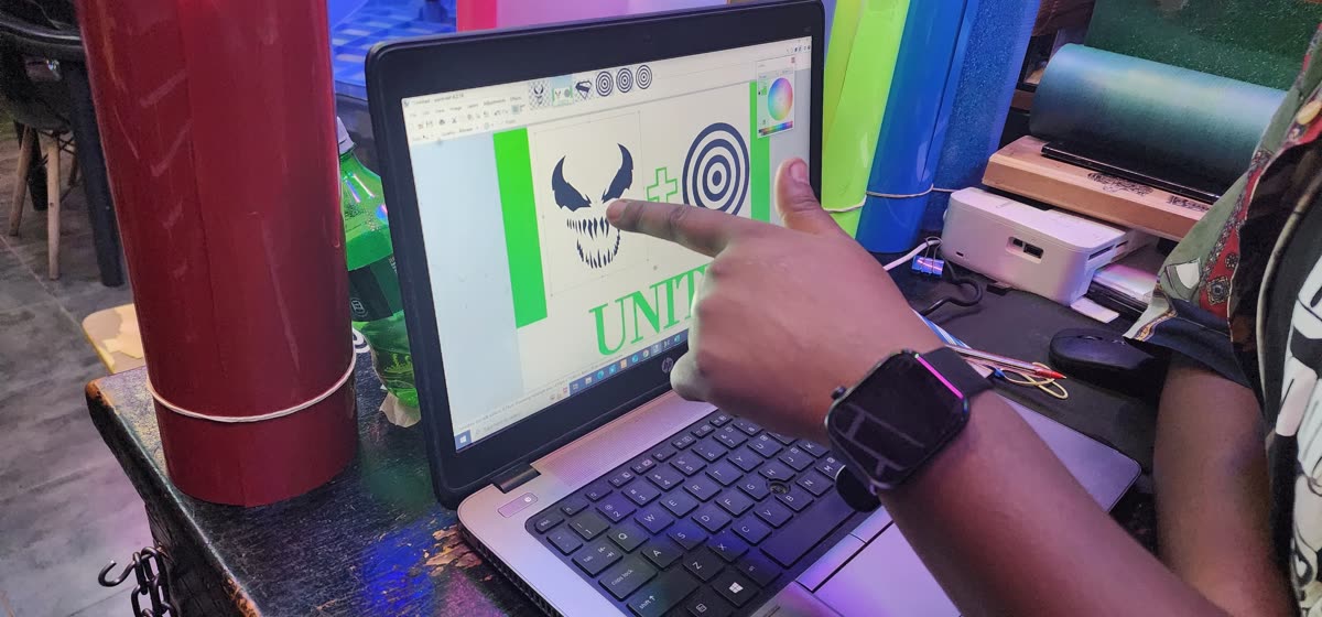 A hand points at a laptop screen showing a black fanged monster-style face graphic, a green plus sign, a black-and-white bullseye target and a green UNITY wordmark in design software. Rolls of red, green, blue and teal vinyl and a small white printer sit beside the laptop.
