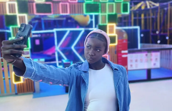 A woman in a denim jacket and pink headband takes a selfie in a neon-lit play arena