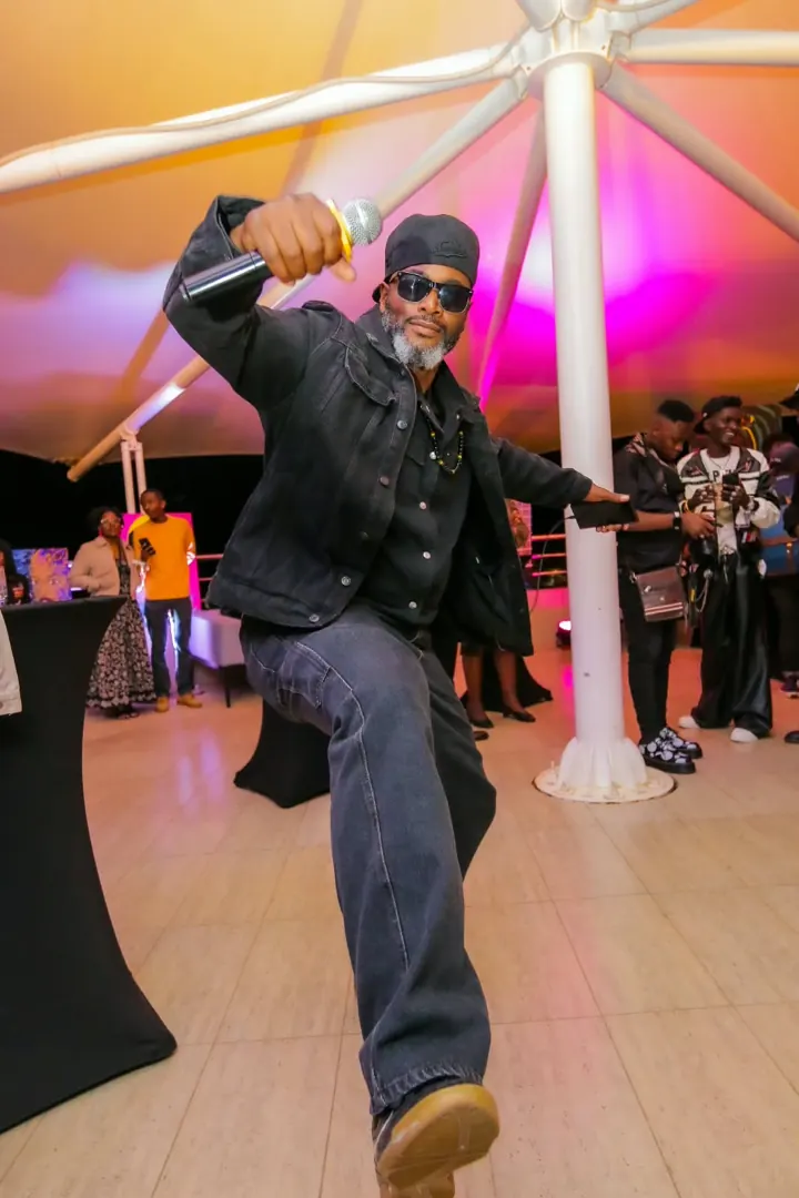 A man in sunglasses and a denim jacket dances with a microphone raised under a white event tent lit pink and orange