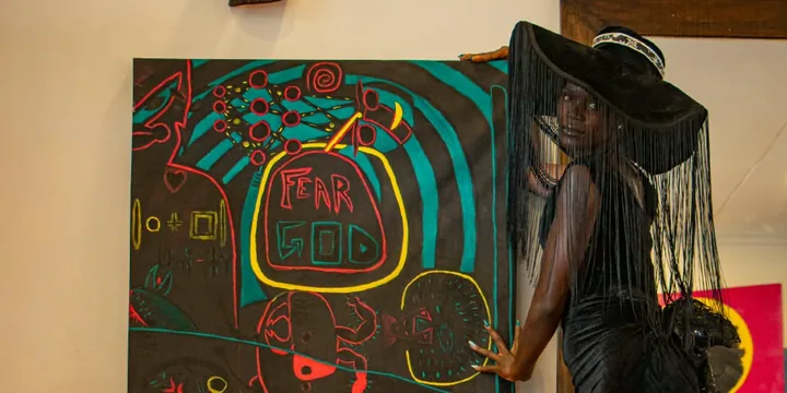 A model in a wide black fringed hat and black gown rests a hand on a large black painting with red, yellow and teal figures and the words FEAR GOD