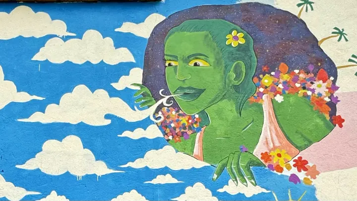 Mural of a green-skinned woman with flowers in her hair against blue sky and white clouds