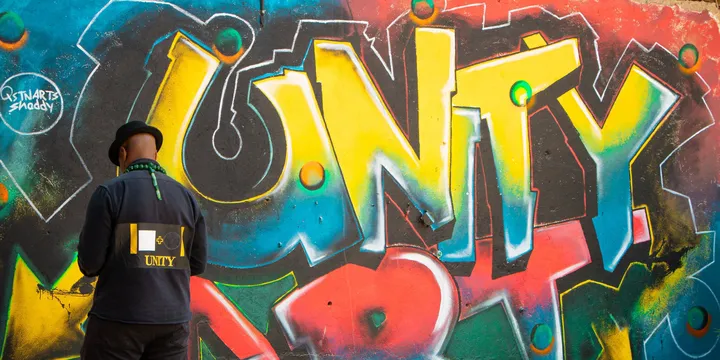 A man in a black UNITY jacket and hat, back to the camera, faces a large graffiti mural reading UNITY ART in yellow, red and blue