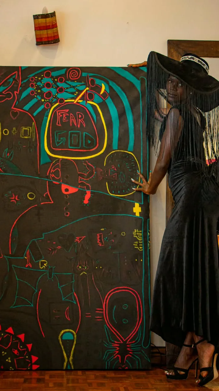The Algorithm of Fate, a large black painting with FEAR GOD at its centre, beside a model in a black fringe hat and gown